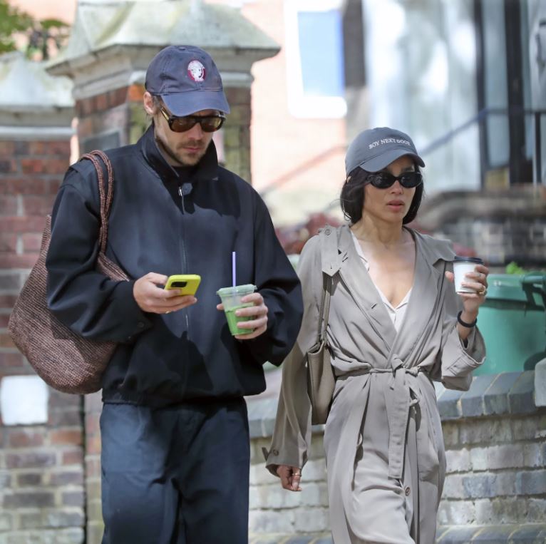 Harry Styles and Zoë Kravitz walking in London wearing coordinated couple outfits with navy caps, trench coat, and casual street style