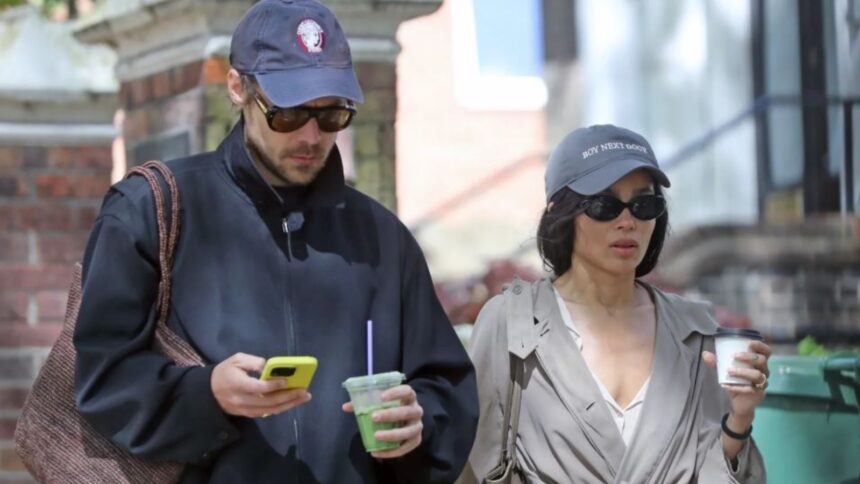 Harry Styles and Zoë Kravitz walking in London wearing coordinated couple outfits with navy caps, trench coat, and casual street style