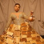 Queen Mercy Atang wearing a dramatic dress made from over 500 loaves of bread at the Africa Magic Viewers' Choice Awards red carpet in Lagos, surrounded by assistants carrying trays of bread.