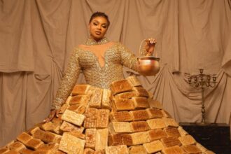 Queen Mercy Atang wearing a dramatic dress made from over 500 loaves of bread at the Africa Magic Viewers' Choice Awards red carpet in Lagos, surrounded by assistants carrying trays of bread.