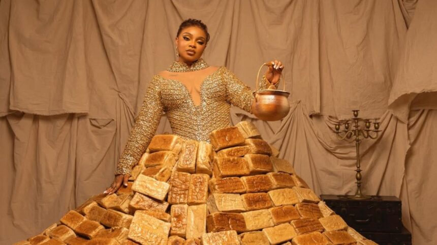 Queen Mercy Atang wearing a dramatic dress made from over 500 loaves of bread at the Africa Magic Viewers' Choice Awards red carpet in Lagos, surrounded by assistants carrying trays of bread.